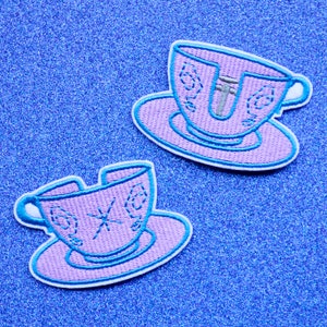 May include: Two purple teacups with blue trim and white saucers. The teacups are cracked and broken. The teacups are on a blue sparkly background.