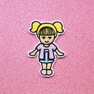May include: An embroidered iron-on patch of a cartoon girl with blonde pigtails, wearing a light purple shirt and a blue skirt. The patch is on a pink glitter background.
