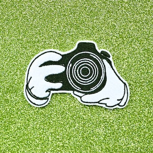 May include: Black and white embroidered patch of a camera being held by two hands. The camera has a large lens with a black and white circular design.