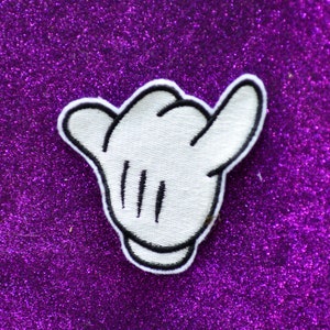 May include: White fabric patch shaped like a hand making the shaka sign, a popular Hawaiian gesture. The patch has black stitching around the edges and black lines for the fingers.