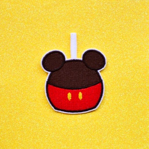 May include: A red and brown felt applique of Mickey Mouse's head with a white stem on a yellow glitter background.