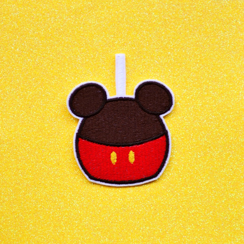 May include: A red and brown felt applique of Mickey Mouse's head with a white stem on a yellow glitter background.