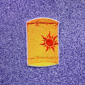 May include: A yellow and orange embroidered patch of a lantern with a sun design. The lantern has a white border and a red sun with orange rays.