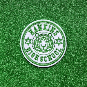 May include: White and green embroidered patch with the text "Hawkins High School" and a tiger head inside a circle. The text "1983" and "1985" are on either side of the tiger head.
