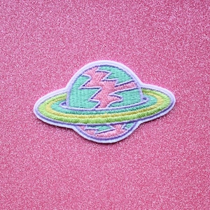May include: A pink, green, and yellow embroidered patch of a planet with a ring around it. The planet has a lightning bolt design.