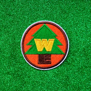 May include: A round, orange and brown embroidered patch with a green pine tree and a yellow "W" in the center. The patch has a white border.
