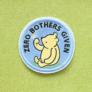 May include: A blue and white embroidered patch with a cartoon bear holding a flower. The text "Zero Bothers Given" is embroidered around the bear.