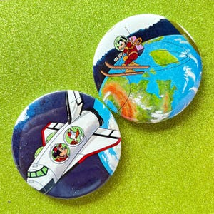 May include: Two circular pinback buttons featuring Disney characters in space. One button shows Goofy skiing in space above Earth. The other button depicts Mickey Mouse and Donald Duck inside a white space shuttle against a dark blue starry sky.