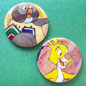 May include: Two circular pin-back buttons featuring cartoon illustrations. One button depicts a grey owl with a yellow beak holding two books. The other button shows a yellow rabbit with a pink nose against a watercolor-style background.