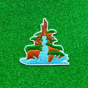 May include: Embroidered patch depicting a waterfall cascading down a brown rock formation with green foliage. The waterfall is blue and white, and the foliage is green. The patch is on a green glitter background.