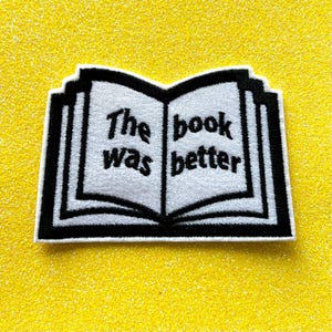 May include: Embroidered patch in the shape of an open book with the text "The book was better" in black lettering on a white background. The patch has a black border and is set against a yellow glitter background.