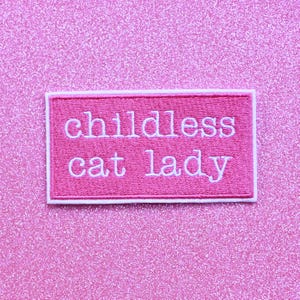 May include: A pink felt patch with white embroidered text that reads "childless cat lady".