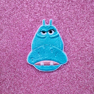 May include: A blue embroidered patch of a cartoon character with a winking eye and a large mouth. The character is shaped like a hippopotamus.