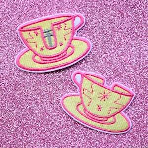 May include: Two yellow and pink embroidered iron-on patches of a teacup and saucer. The teacups are cracked and have a star design.