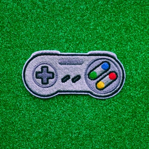 May include: A gray and white video game controller with colored buttons. The controller is embroidered on a green sparkly background.