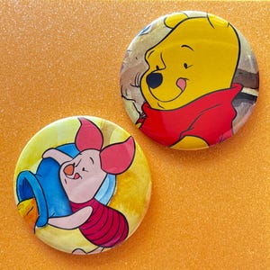 May include: Two circular pin-back buttons featuring classic cartoon characters. One button displays a yellow bear in a red shirt, while the other button shows a small pink piglet wearing a striped shirt and holding a blue honey pot.