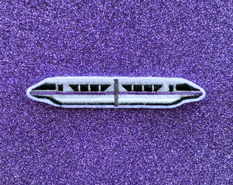 May include: White and purple embroidered patch of a monorail train on a purple glitter background.