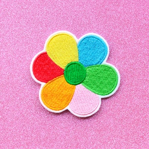 May include: A colorful embroidered flower patch with six petals in shades of red, orange, yellow, green, blue, and pink. The center of the flower is green.