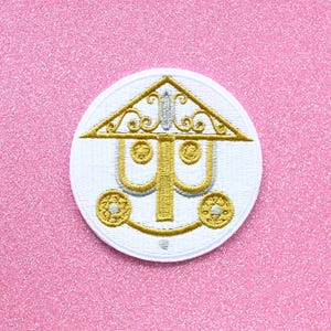 May include: A white embroidered patch with a gold and silver design of a carriage with a pointed roof. The carriage has two wheels and a decorative top.