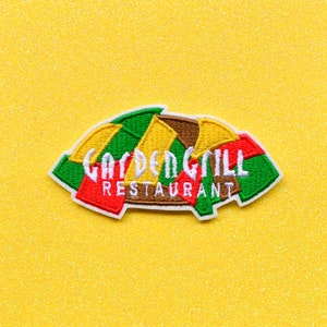 May include: A colourful embroidered patch with the text "GARDEN GRILL RESTAURANT" in white lettering. The patch is shaped like a half-circle and features a green, yellow, red and brown design.