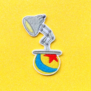 May include: A gray and white embroidered patch of a lamp with a yellow light and a red star on a blue and yellow ball.