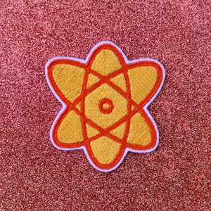 May include: A yellow and orange embroidered atom patch with a white outline. The patch is on a red glitter background.