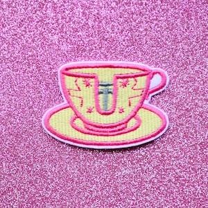 May include: A yellow teacup and saucer with a pink outline and a gray cross inside. The teacup has a white rim and a white interior. The saucer has a white rim. The teacup and saucer are on a pink glitter background.