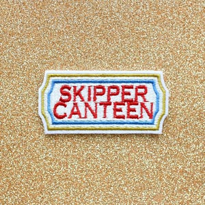 May include: Embroidered patch with the words "SKIPPER CANTEEN" in red, outlined in blue, and framed by a gold and white border. The patch is rectangular with rounded corners, set against a gold glitter background.