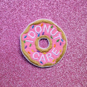 May include: Pink donut-shaped embroidered patch with the text "Donut Care" in white lettering. The donut has a brown outline and colorful sprinkles.