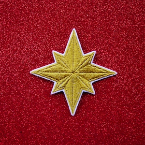 May include: A gold star-shaped patch with white stitching on a red glitter background.