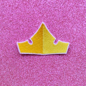 May include: A yellow felt crown with white stitching on a pink glitter background.