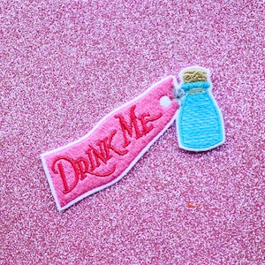 May include: Pink fabric tag with white trim and the words "Drink Me" embroidered in red. A blue bottle with a cork top is attached to the tag.