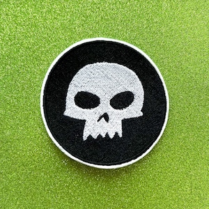 May include: Round black and white embroidered patch featuring a skull design. The skull is white with black eye sockets and a jagged jawline, set against a black background. The patch has a white border and is on a green surface.