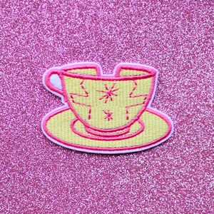 May include: An embroidered patch shaped like a yellow teacup on a saucer with a bright pink stitched border. The teacup features a jagged crack design and two small star-shaped accents in pink thread, with a white fabric backing visible at the edges.