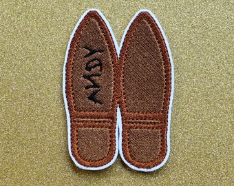 Puede incluir: Un par de zapatillas de fieltro bordadas de color marrón con la palabra "Yay" bordada en una zapatilla.