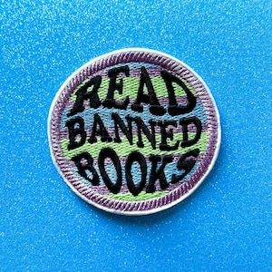 May include: A circular embroidered patch featuring the text "READ BANNED BOOKS" in black, with green, blue, and purple accents. The patch has a white border and is set against a bright blue background.