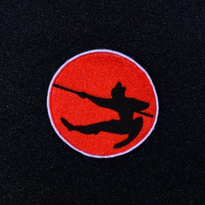 Peut inclure: Patch brodé rouge avec un contour blanc représentant une silhouette noire d'une personne en position de combat d'arts martiaux tenant un bâton.