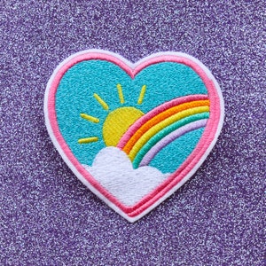 May include: A pink heart-shaped patch with an embroidered design of a blue sky, a yellow sun, and a rainbow with pink, orange, yellow, green, and purple stripes.
