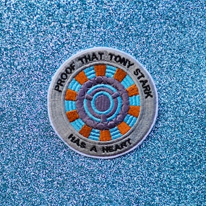 May include: A round, embroidered patch with a gray background and a blue and orange design in the center. The text "Proof that Tony Stark has a heart" is written around the edge of the patch.