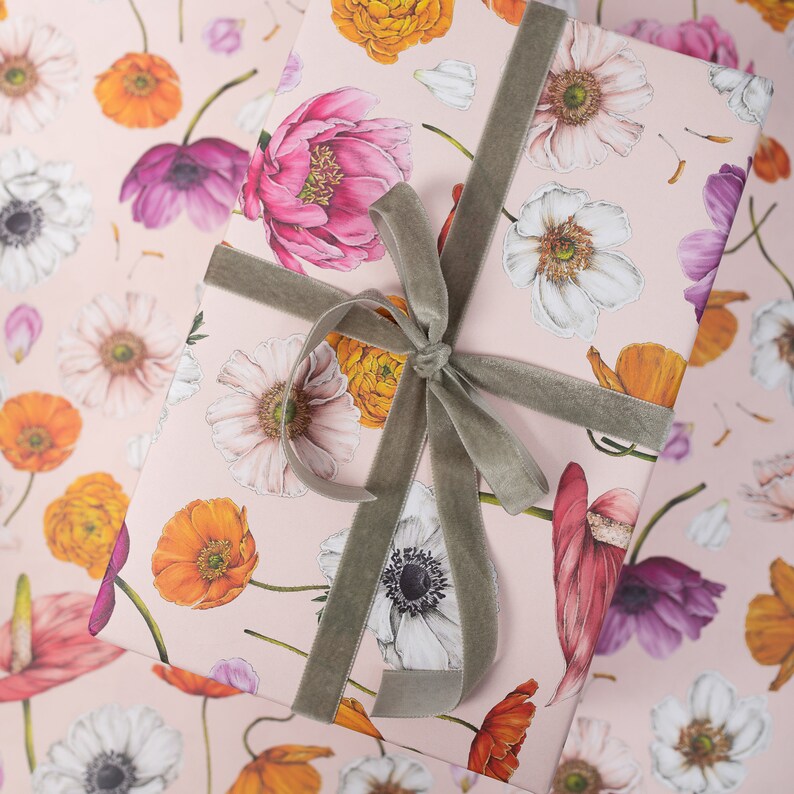 May include: A rectangular gift box wrapped in light pink paper featuring a repeating floral pattern of orange, pink, white, and purple flowers. The box is tied with a soft, grey velvet ribbon that is knotted in a bow on the top surface.
