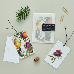 May include: Gift tags with botanical illustrations of pears, flowers, and pinecones. One tag has "TO: FROM:" text. A package of tags is labeled "GIFT TAGS 6 pk with twine". Gold paper clips and a sprig of greenery are also present.