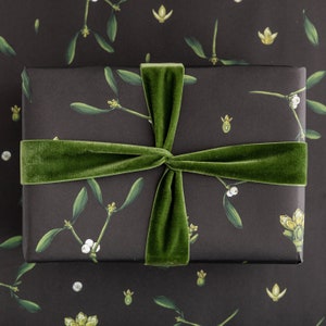 May include: A wrapped gift box featuring a dark background with a botanical mistletoe pattern. The box is tied with a wide, green velvet ribbon. The mistletoe design includes green leaves, white berries, and yellow flower details.