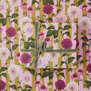 May include: A wrapped gift box with a green velvet ribbon and bow sits atop floral patterned wrapping paper. The paper features pink stripes and a repeating pattern of white and magenta dahlias with green leaves.
