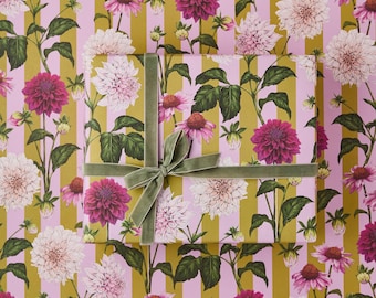 May include: A wrapped gift box with a green velvet ribbon and bow sits atop floral patterned wrapping paper. The paper features pink stripes and a repeating pattern of white and magenta dahlias with green leaves.