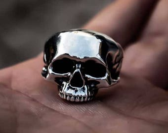 May include: A silver skull ring with a detailed design. The ring is on a person's hand.