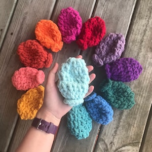 May include: A collection of 12 colorful crocheted bottle cozies in various shades of red, orange, yellow, green, blue, purple, and pink. The cozies are arranged in a circle on a wooden surface.