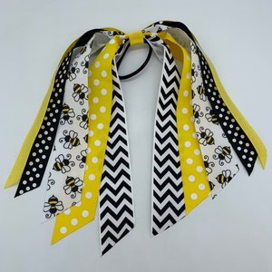 May include: A set of yellow, black and white hair ties with a bee pattern. The hair ties are made of ribbon and have a chevron, polka dot and bee pattern.