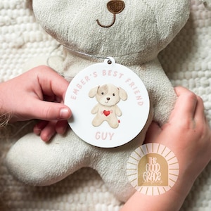 May include: A beige plush dog toy with a brown embroidered nose and eyes, featuring a circular white tag attached by a silver string. The tag displays a graphic of a dog with a red heart and the text Ember's Best Friend Guy in pink letters.