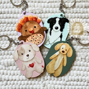 May include: Four oval keychains featuring watercolor-style illustrations: a brown turkey with orange feathers, a black and white dog, a light brown puppy with a red heart, and a tan plush toy. Each keychain includes a metal ring and chain.