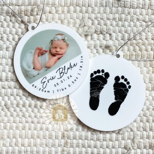 May include: Two round white ornaments with silver hanging strings. One ornament features a photograph of a newborn baby wearing a floral headband, with the text Evie Blake, 05.01.24, 09:20 am, 7 lbs 0.5 oz, and 18.5 in. The other ornament displays black baby footprints.